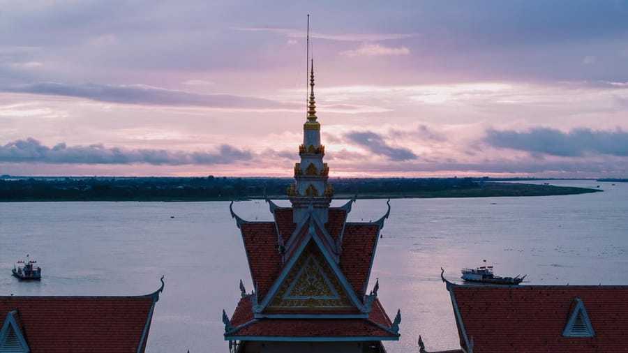 A gold-tipped Khmer temple spire and tiled roofline overlooking a wide river at dusk, with boats passing on the water.