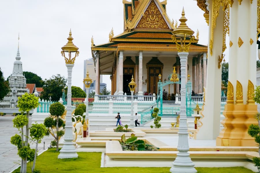 The gilded pavilion of Phnom Penh’s Royal Palace grounds, seen past ornate lampposts and a manicured lawn with visitors walking by.