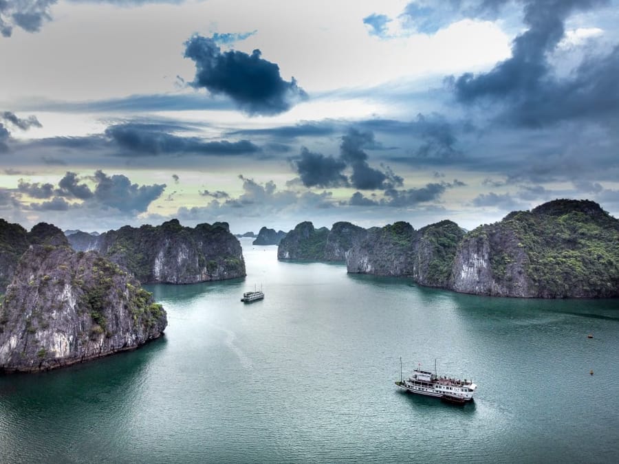 Two tour boats cruising past limestone karst islands in Hạ Long Bay under a dramatic, cloud-heavy sky.