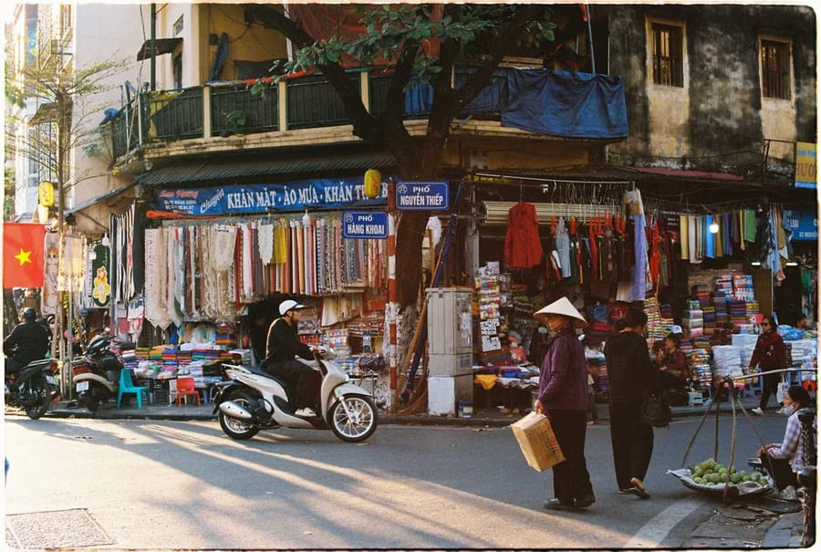 A street corner in Hanoi’s Old Quarter, with fabric stalls, a passing scooter, and a woman in a conical hat carrying a box.