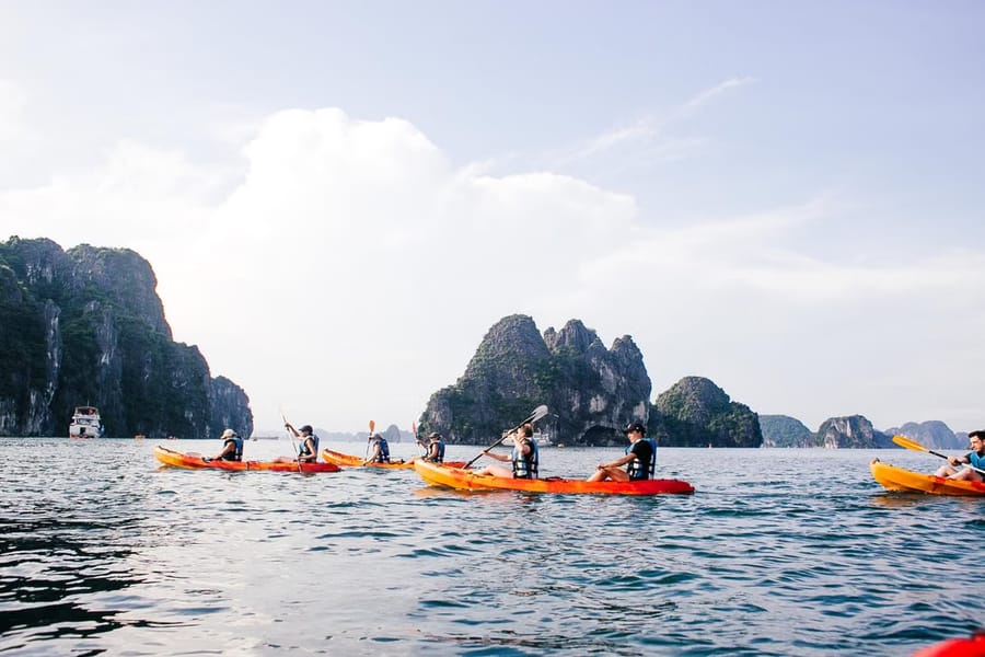 Kayakers in orange kayaks paddle across calm water among the limestone karsts of Hạ Long Bay.