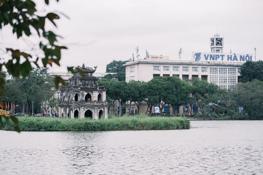The Turtle Tower stands on its small islet in Hoàn Kiếm Lake, Hanoi, with a clock-tower building visible on the far shore.