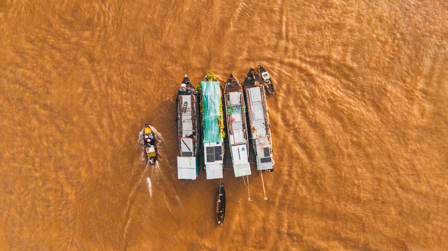Seen from directly above, a row of moored cargo boats sits on the brown Hậu River at Cái Răng floating market while a small sampan motors past.