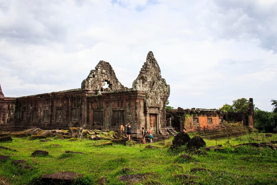Visitors walk past the crumbling sandstone gables of the Wat Phou temple ruins in southern Laos, with green grass and scattered blocks in front.
