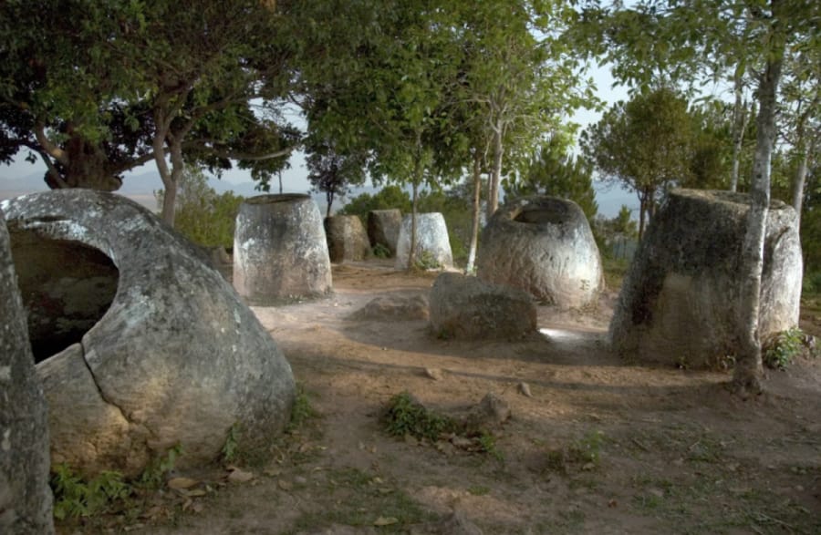 Weathered stone jars, some taller than a person and one cracked open, stand among trees on a dry hillside at the Plain of Jars in Laos.