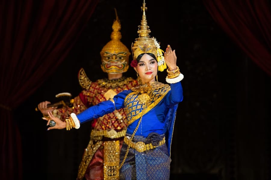 A Khmer classical dancer in a gold crown and blue silk holds a graceful pose on stage, with a masked giant character behind her.